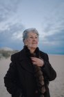 Elderly female tourist with gray hair in warm casual outfit looking away while relaxing on sandy beach against cloudy evening sky — Stock Photo