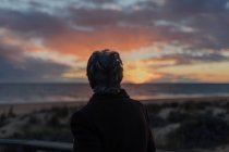 Back view of elderly female traveler in casual clothes standing on wooden pier on sandy beach and enjoying seascape at sunset — Fotografia de Stock