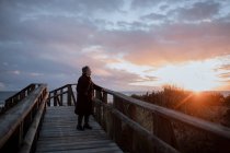 Vista laterale di anziano viaggiatore femminile in abiti casual in piedi su un molo di legno sulla spiaggia di sabbia e godersi il paesaggio marino al tramonto — Foto stock