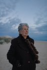 Elderly female tourist with gray hair in warm casual outfit looking away while relaxing on sandy beach against cloudy evening sky — Stock Photo