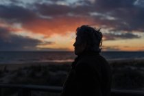 Seitenansicht einer älteren Reisenden in Freizeitkleidung, die auf einem hölzernen Pier am Sandstrand steht und das Meer bei Sonnenuntergang genießt — Stockfoto