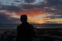 Rückansicht einer älteren Reisenden in Freizeitkleidung, die auf einem Holzsteg am Sandstrand steht und das Meer bei Sonnenuntergang genießt — Stockfoto