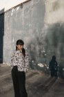 Pensiva jovem senhora étnica com longos cabelos escuros em vestido casual em pé na rua perto de casas antigas e olhando para longe no dia ensolarado — Fotografia de Stock