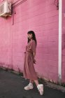 Вид сбоку на стильную молодую этническую женщину с длинными темными волосами в модном платье, стоящую напротив розовой стены на улице и задумчиво смотрящую в сторону — стоковое фото