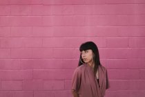 Вид сбоку на стильную молодую этническую женщину с длинными темными волосами в модном платье, стоящую напротив розовой стены на улице и задумчиво смотрящую в сторону — стоковое фото