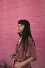 Seitenansicht einer stilvollen jungen ethnischen Frau mit langen dunklen Haaren im trendigen Kleid, die an einer rosa Wand auf der Straße steht und nachdenklich wegschaut — Stockfoto