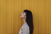 Side view of stylish young ethnic female with long dark hair in trendy dress standing against yellow wall on street and closed eyes thoughtfully — Stock Photo