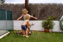 Side view of cheerful shirtless little boy pouring water from hose on sister in swimsuit while playing together in yard on summer day — Stock Photo