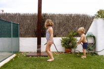 Seitenansicht des fröhlichen, hemdlosen kleinen Jungen, der an einem Sommertag im Hof zusammen mit seiner Schwester im Badeanzug Wasser aus dem Schlauch gießt — Stockfoto