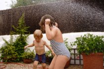 Vue latérale de joyeux petit garçon torse nu versant de l'eau du tuyau sur la soeur en maillot de bain tout en jouant ensemble dans la cour le jour de l'été — Photo de stock