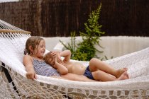 Cute barefooted little sister and brother hugging while lying in hammock and having fun during summer holidays — Stock Photo