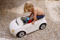Vista laterale di carino allegro bambino con capelli biondi equitazione giocattolo auto mentre posa in cortile nella soleggiata giornata estiva — Foto stock