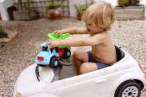Side view of cute cheerful little boy with blond hair riding toy car while laying in yard on sunny summer day — Stock Photo