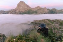 From above back view of anonymous male tourist taking photo of mount in fog on camera under cloudy sky — Stock Photo