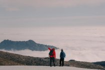 Vista posterior de exploradores anónimos en ropa interior cálida con mochilas y bastones de trekking parados en el pico nevado de la montaña escondidos bajo densas nubes - foto de stock