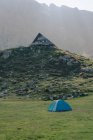 Tente de camping située sur une prairie herbeuse près d'une colline avec maison résidentielle au sommet entourée de montagnes massives par une journée ensoleillée — Photo de stock