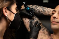 Female tattoo master in mask and with machine making tattoo on arm of male client in dark salon — Stock Photo