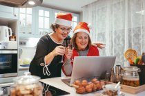 Femmes mûres homosexuelles sincères chapeaux Santa avec champagne regarder netbook avec chat vidéo pendant les vacances du Nouvel An dans la cuisine — Photo de stock