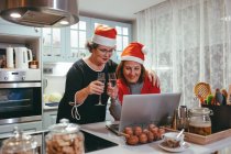 Femmes mûres homosexuelles sincères chapeaux Santa avec champagne regarder netbook avec chat vidéo pendant les vacances du Nouvel An dans la cuisine — Photo de stock