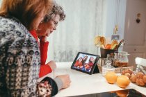 Вид збоку усміхнених гомосексуальних літніх жінок, які дивляться планшет із щасливими дітьми та матерями під час відеочату вдома — стокове фото