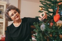 Donna di mezza età concentrata in occhiali con testa inclinata che decora l'abete prima di vacanza di anno nuovo in casa — Foto stock