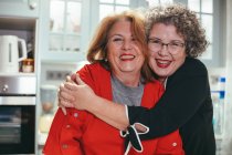 Frohe lesbische Frau mittleren Alters umarmt lächelnde Geliebte im Haus und schaut in die Kamera — Stockfoto