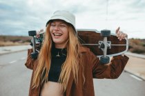 Весела молода жінка тисячоліття в стильному вбранні і капелюсі сміється з закритими очима, стоячи на асфальтній дорозі зі скейтбордом за головою після їзди — стокове фото