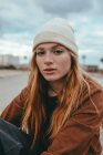 Selbstbewusste junge Frau mit langen Ingwerhaaren steht an bewölkten Tagen auf der Straße und blickt in die Kamera — Stockfoto