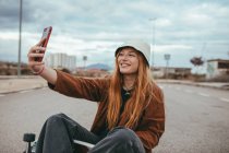 Счастливая девушка-подросток с длинными рыжими волосами в модном наряде и шляпе сидит на скейтборде и улыбается, делая селфи на мобильном телефоне — стоковое фото
