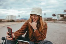 Adolescente mujer feliz con el pelo largo y rojo en traje de moda y sombrero sentado en el monopatín y sonriendo mientras toma selfie en el teléfono móvil - foto de stock