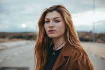 Giovane donna fiduciosa con lunghi capelli rossi in piedi sulla strada il giorno nuvoloso e guardando la fotocamera — Foto stock