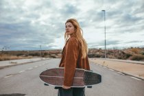 Vista lateral da jovem patinadora com longos cabelos loiros em roupas da moda em pé na estrada de asfalto com skate cruzador na mão contra o céu nublado no campo — Fotografia de Stock