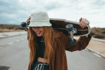 Allegro giovane donna millenaria in abito elegante e cappello ridere ad occhi chiusi mentre in piedi su strada asfaltata con skateboard dietro la testa dopo la guida — Foto stock