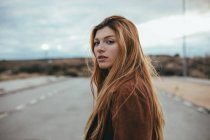 Vue latérale d'une jeune femme confiante avec de longs cheveux roux debout sur la rue par temps nuageux et regardant la caméra — Photo de stock