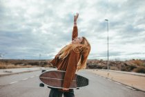 Side view of young female skater with long blond hair in trendy outfit standing on asphalt road with cruiser skateboard in hand against cloudy sky in countryside — Stock Photo