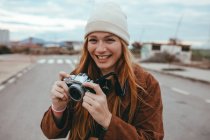 Позитивна молода леді з довгим імбирним волоссям в стильному одязі і капелюсі посміхається, стоячи на дорозі з старовинною фотокамерою в руці під час поїздки в сільській місцевості — стокове фото