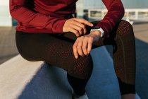 Schnuppern anonyme Sportlerin in stylischer Sportbekleidung sitzt am Rand und überprüft den Puls auf Fitness-Tracker, während sie während des Outdoor-Trainings in der Stadt ruht — Stockfoto