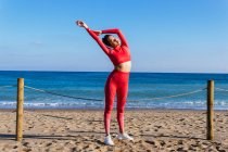 Auto-assegurada jovem atleta étnica feminina em sportswear vermelho em pé na praia de areia perto do oceano ondulado e esticando os braços antes do treinamento — Fotografia de Stock
