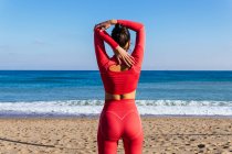 Back view female athlete in red sportswear standing on sandy beach near wavy ocean and stretching arms before training — Stock Photo