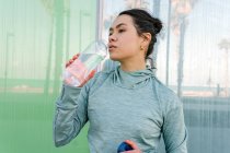 Спокійна молода етнічна жінка-спортсменка з темним волоссям у спортивному одязі п'є воду з пляшки під час тренування на відкритому повітрі на морі в сонячний день — стокове фото