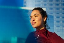 Confident young ethnic female athlete in stylish sportswear looking away while standing against modern glass building on city street — Stock Photo
