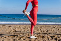 Vue latérale d'une jeune dame en tenue ethnique méconnaissable en vêtements de sport et baskets étirant les jambes tout en se tenant debout sur la plage de sable fin pendant l'entraînement en plein air — Photo de stock