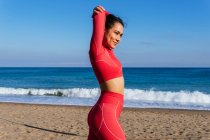 Самозабезпечена молода етнічна жінка-спортсменка в червоному спортивному одязі, що стоїть на піщаному пляжі біля хвилястого океану і розтягує руки перед тренуванням — стокове фото