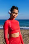 Молода іспанська спортсменка в навушниках з темним волоссям в червоному костюмі посміхається і дивиться на камеру під час тренування на піщаному пляжі проти безхмарного блакитного неба. — стокове фото