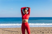 Самоуверенная молодая спортсменка в красной спортивной одежде стоит на песчаном пляже возле волнистого океана и вытягивает руки перед тренировкой — стоковое фото
