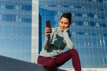 Glückliche junge hispanische Dame in Sportkleidung, die lächelt und ihr Handy benutzt, während sie sich nach dem Outdoor-Training an einem sonnigen Tag auf einer Bank in der Nähe eines modernen Gebäudes ausruht — Stockfoto