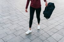 Crop fit femminile in abito sportivo alla moda e scarpe da ginnastica che trasportano borsetta mentre si cammina sulla piazza pavimentata della città — Foto stock