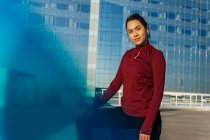 Jovem atleta étnica confiante em roupas esportivas elegantes olhando para longe enquanto estava contra o edifício de vidro moderno na rua da cidade — Fotografia de Stock