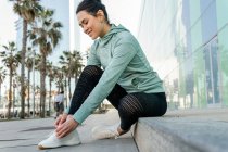 Side view of smiling young ethnic female athlete in sportswear tying sneakers during outdoor workout on city embankment on sunny day — Stock Photo
