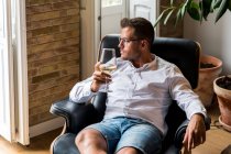 Homme pensif reposant dans un fauteuil en cuir confortable avec un verre de vin blanc et regardant loin dans les pensées — Photo de stock